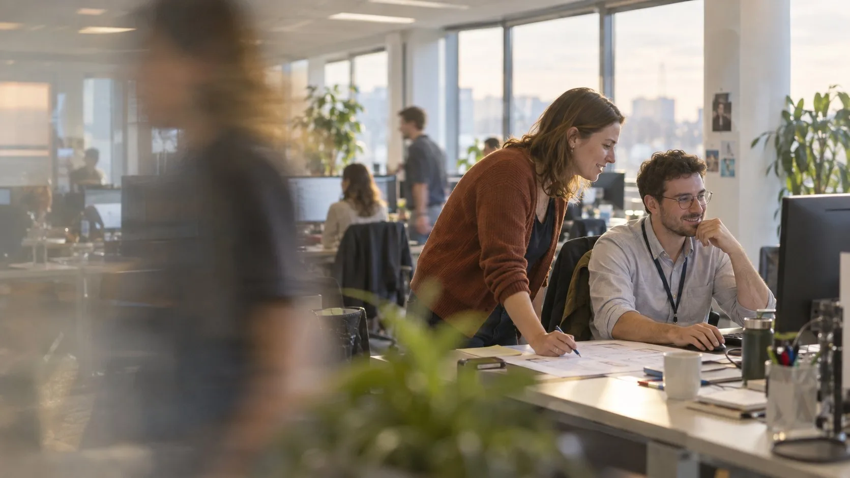 A young consultant leans over a printed process map and smiles with a finance manager at his monitor in a bright open-plan office, with colleagues passing as blurred figures